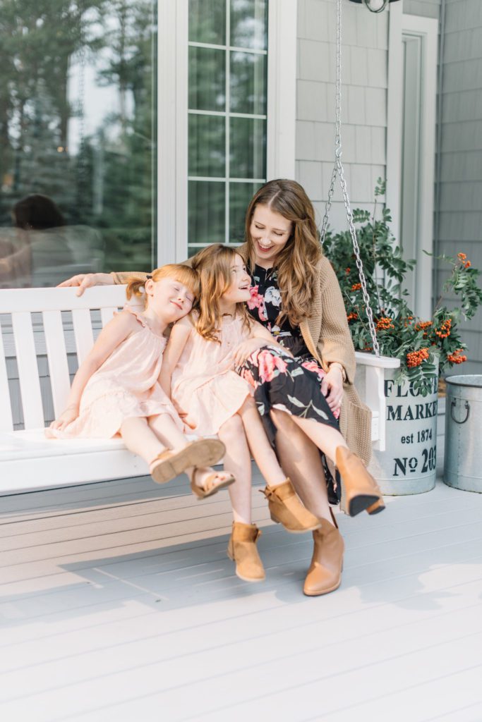 A mom sits with 2 little girls on a white porch swing