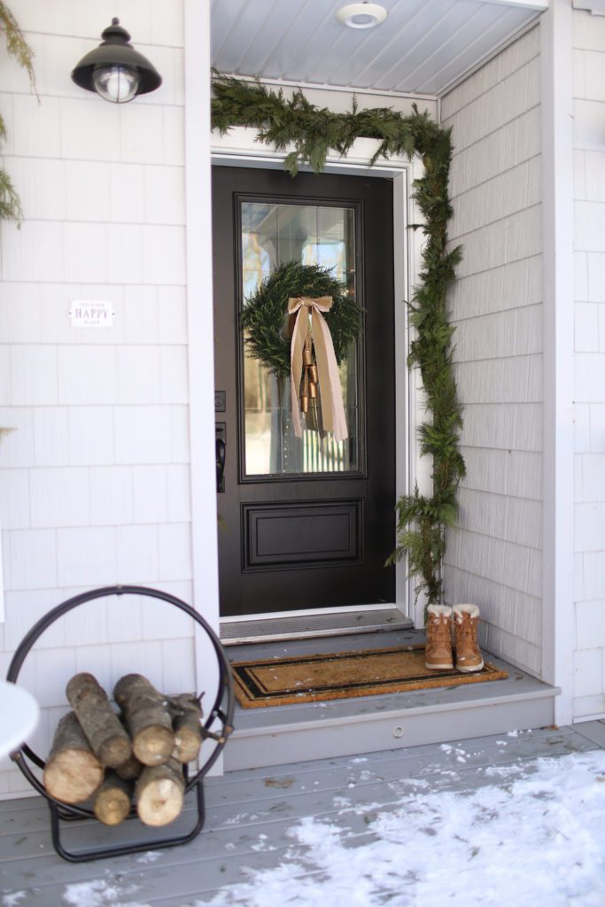 A black front door with a Christmas wreath and a round log holder 
