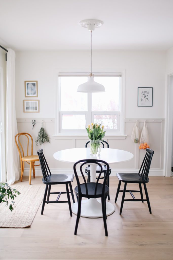 A small dining space with a round white table, black chairs and a hanging pendant light with beige panelling and a window in the wall behind.