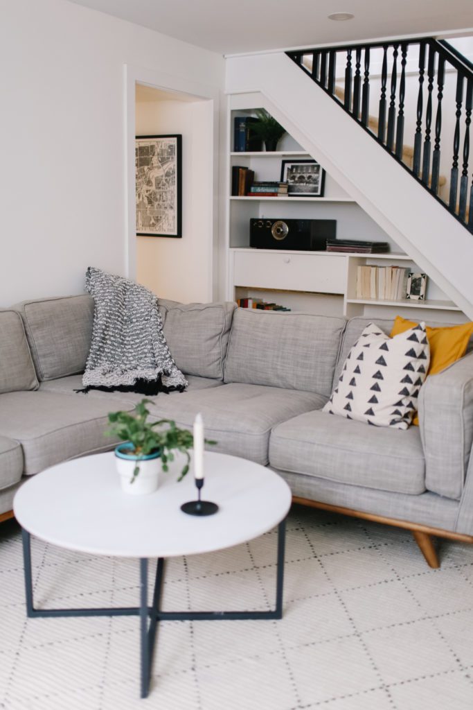 A cozy basement setup with a grey sectional, white coffee table, pillow and throws.